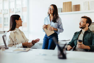 a woman leans against a desk while talked to a man and a woman in chairs in front of her