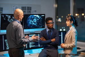 a man leans against a desk with his arms crossed talking to two people in front of him